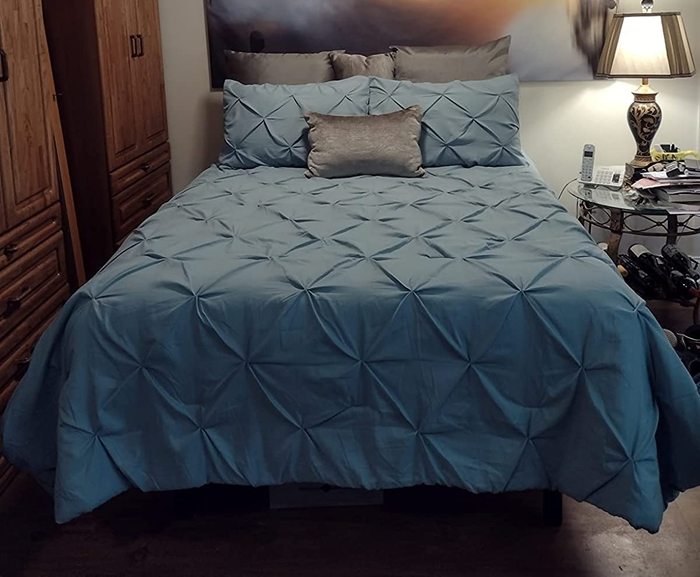 A reviewer image showing a teal pleated full-size comforter set with matching shams, neatly arranged on a bed next to a wooden closet