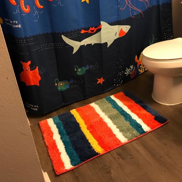 Reviewer photo of a multi-colored striped bath mat placed in front of a shower curtain featuring a shark design