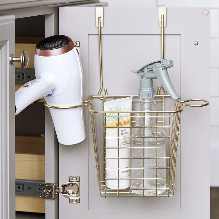 An open bathroom cabinet door featuring a basket-like over-the-door organizer in gold, filled with hair tools.
