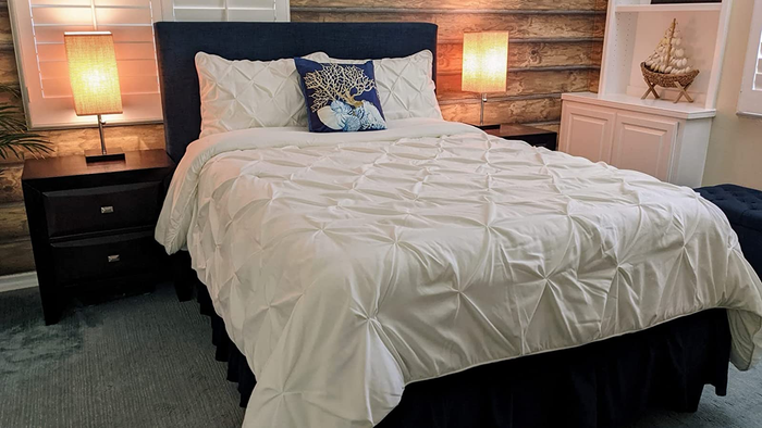 A reviewer image showing a white pleated full-size comforter with matching pillowcases, placed on a wooden bed frame with matching side tables.