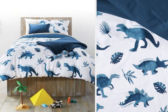 A blue and white dinosaur watercolor-patterned comforter with a matching pillow, next to a navy blue pillow and a solid blue throw blanket with pom-poms, placed on a wooden bedframe. A close-up view of the comforter shows a navy blue underside.