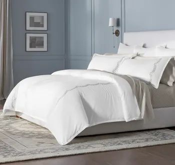 White bedding featuring gray scalloped vine embroidery on the duvet and coordinating white pillows atop a white bed frame.