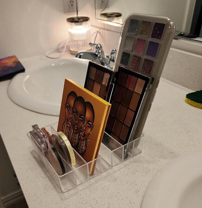 A clear organizer on a bathroom countertop neatly displays an assortment of makeup palettes and compacts, showcasing various shades perfect for any beauty routine.