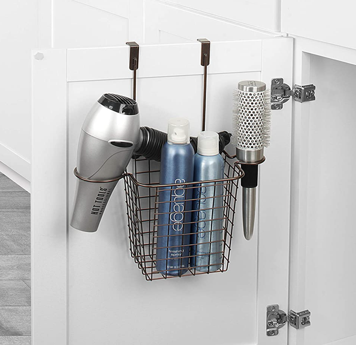 An open bathroom cabinet door displaying a basket-like over-the-door organizer in bronze, filled with hair tools.