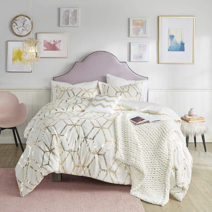 A white and gold sparkly geometric comforter with matching pillowcases and a white knit blanket, elegantly draped over a purple headboard for a chic and modern touch.