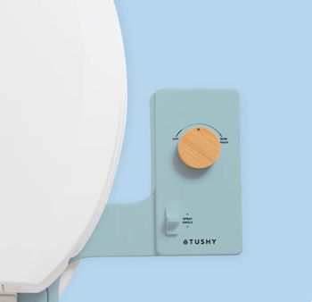 A close-up shot of a TUSHY bidet attachment featuring a wooden control knob, installed on a toilet seat, highlighted in a bathroom accessories shopping guide.