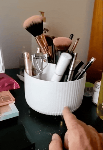 A makeup organizer featuring brushes, lipsticks, mascara, and an eyelash curler on a vanity table, with someone's hand rotating it.