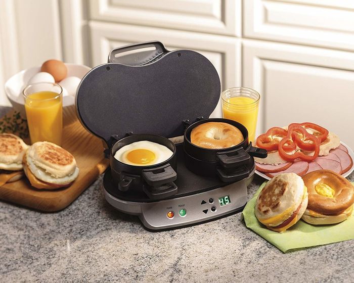Two sandwiches being cooked in the maker, surrounded by ingredients like bread, bagels, eggs, ham, and more.