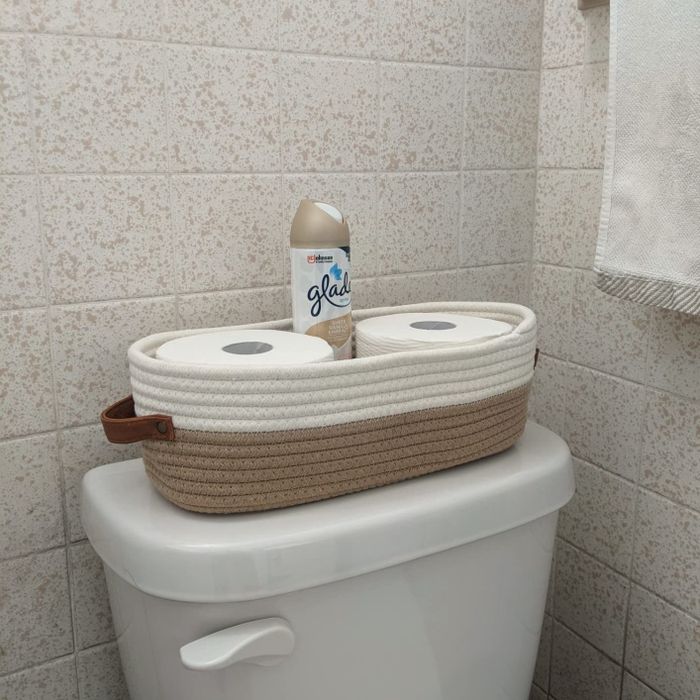 A woven basket containing toilet paper, with an air freshener placed on top of a toilet tank.