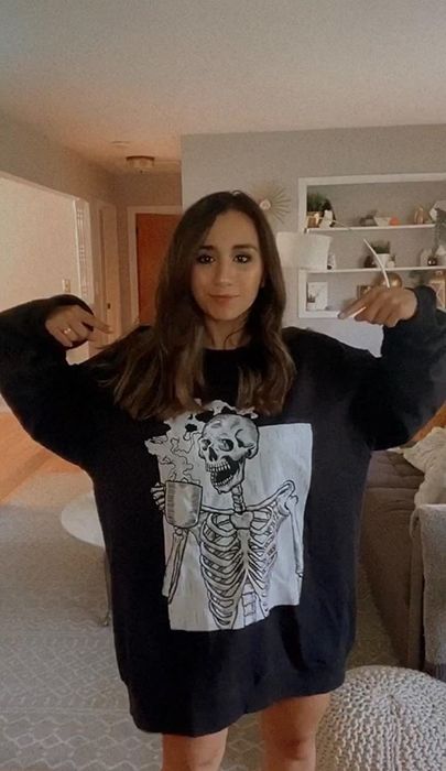 a reviewer in an oversized black sweatshirt featuring a white graphic of a skeleton drinking coffee
