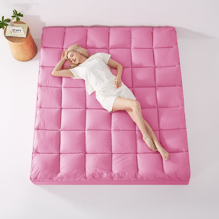 A person lounging on a large, plush pink mattress in casual sleepwear. A small side table with a plant sits nearby