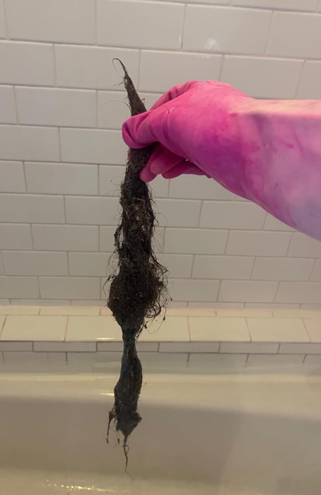 A hand wearing a pink glove holding a large ball of hair extracted from a bathtub drain, highlighting a typical plumbing issue.