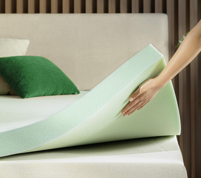 A hand lifts and touches a green tea-infused memory foam mattress topper on a bed