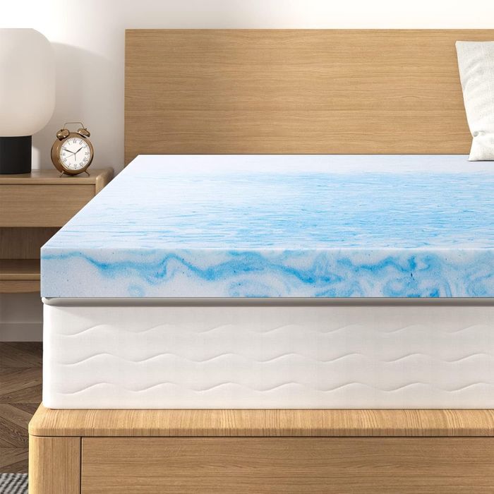 A memory foam mattress topper with a blue swirl pattern, placed on a bed frame in a sleek, modern bedroom. An alarm clock and a lamp are set on the nightstand.