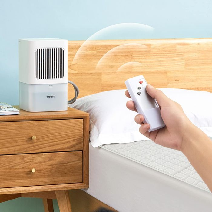 A person adjusting the settings of a compact air purifier using a remote on a bedside table in a contemporary bedroom