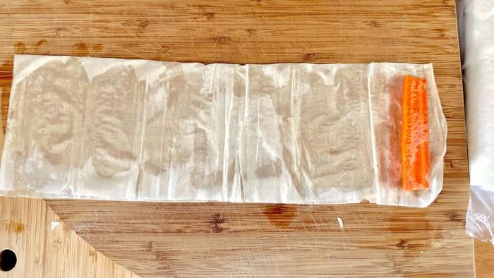 A carrot stick rests at one end of a buttered and folded sheet of phyllo dough.