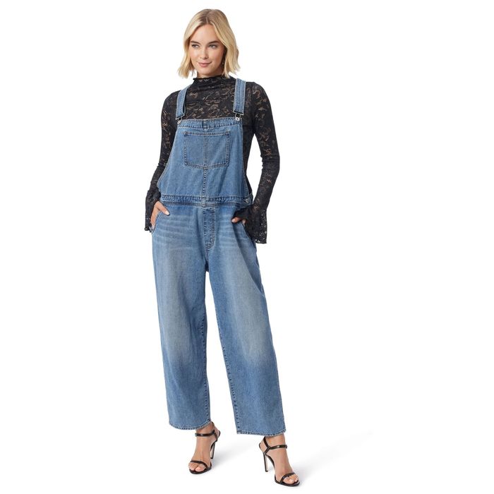 A woman posing in denim overalls and a black lace top, styled with strappy high heels, exuding effortless charm.