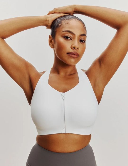 A person modeling a white sports bra with a front zipper, raising their arms confidently.