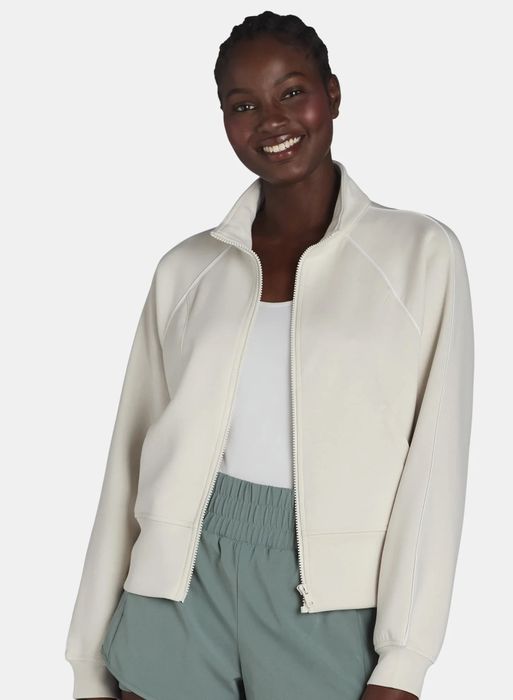 A woman smiling, dressed in a casual zip-up jacket over a tank top and shorts, looking relaxed and comfortable.