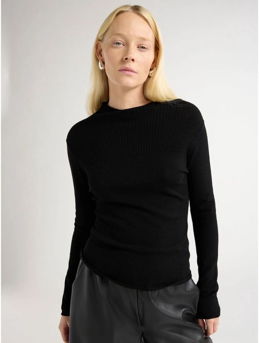 Person styled in a black ribbed sweater and dark fitted pants, posing in a minimalist setting
