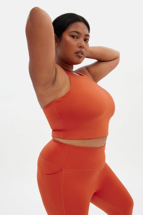 A model in vibrant orange activewear, performing a stretch while wearing a sleeveless crop tank sports bra and leggings
