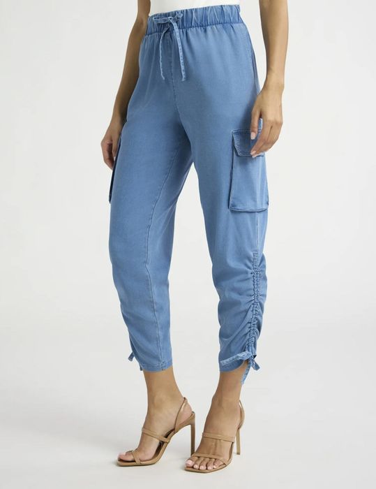 A woman is seen wearing blue jogger pants with cargo pockets and ruched detailing, teamed with a simple white top and tan sandals, creating a casual yet fashionable look.