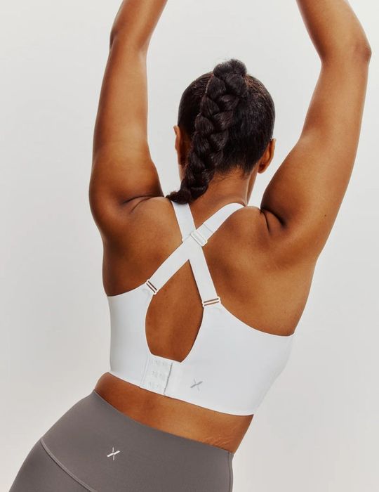 The same model is now wearing a white sports bra with crisscrossed straps at the back and gray leggings, hands lifted above the head.