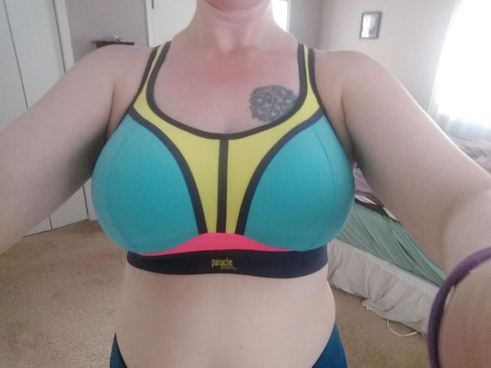 A person wearing a sporty, color-blocked bra, highlighting the front design details. The background shows a cozy home environment with bedroom furniture in view.