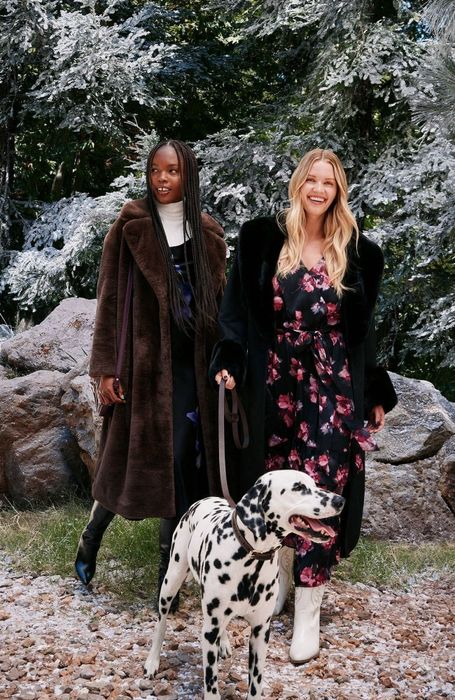 Two people stand outdoors in stylish long coats, one featuring a floral print, accompanied by a Dalmatian dog, with snow-covered trees in the background.