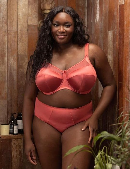 A model in a stylish coral lingerie set, striking a pose in a rustic ambiance with skincare products neatly arranged in the background.
