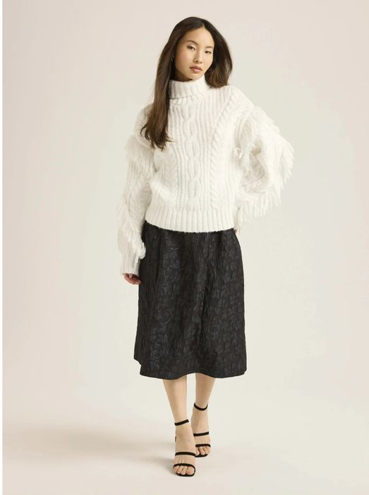 Person wearing a chunky white turtleneck sweater paired with a black knee-length skirt, standing with a neutral expression