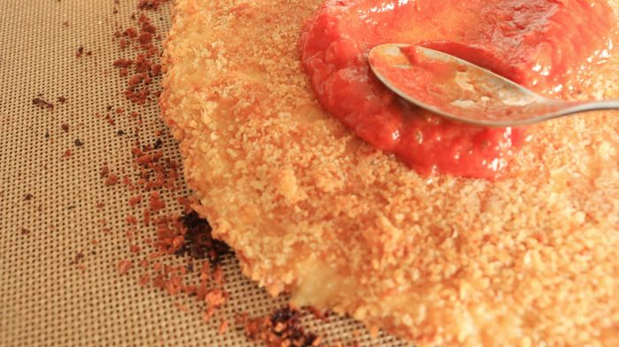 A spoon spreading a layer of tomato sauce onto the chicken crust.