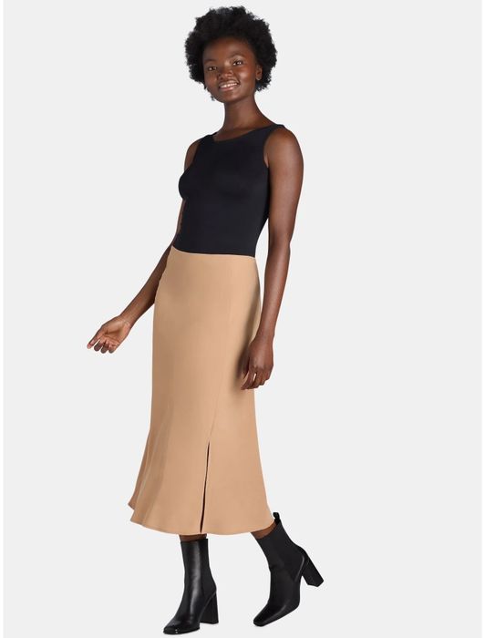 Woman wearing a sleeveless black top, beige midi skirt, and black ankle boots, striking a confident pose in a fashion editorial