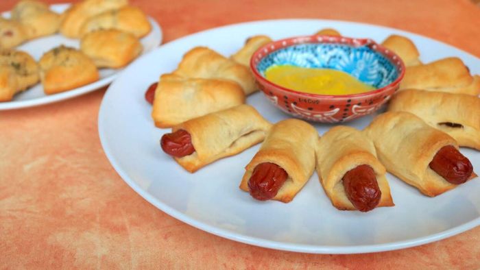 Mini hot dogs wrapped in crescent dough served on a plate with a small dish of mustard in the center.
