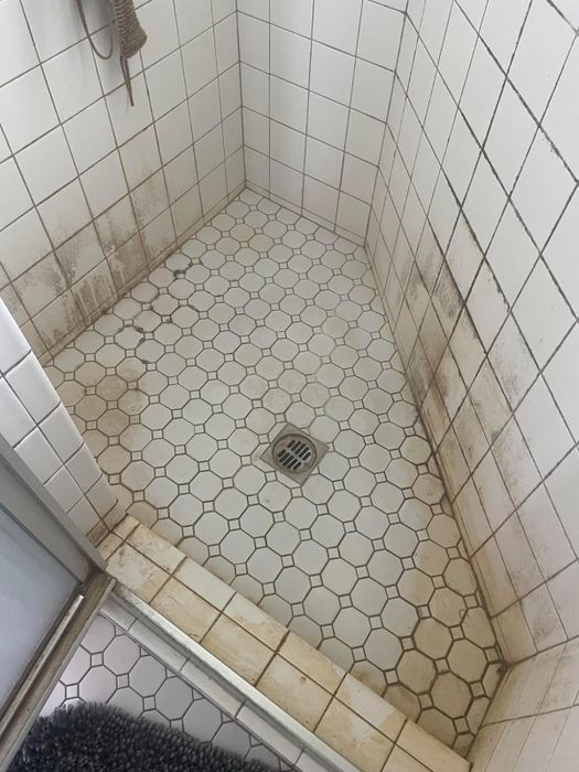 A shower stall with white hexagonal floor tiles and square wall tiles, showing noticeable stains and mold buildup. No people are visible