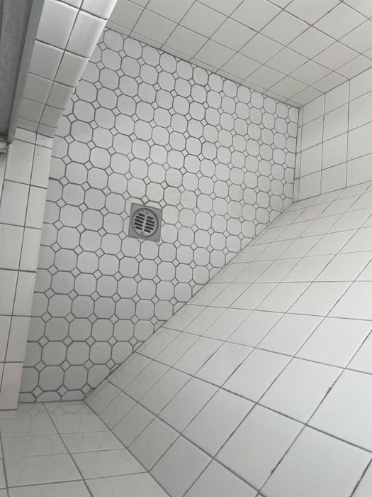 A bathroom shower space with white tiled walls and floor, featuring hexagonal tiles on the floor and a drain positioned in the center