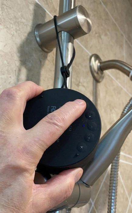 A hand reaching up to adjust a black, round Bluetooth shower speaker hanging from the shower head