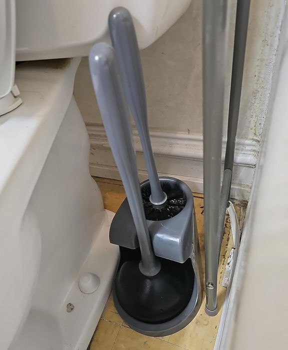 A gray toilet cleaning brush and plunger in a holder placed between a toilet and a wall.