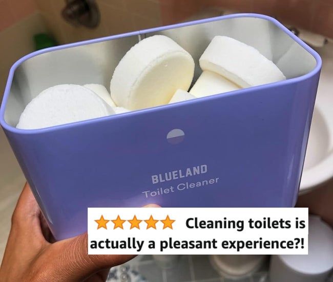A hand holding a Blueland Toilet Cleaner container with white tablet cleaners inside, placed on a bathroom floor