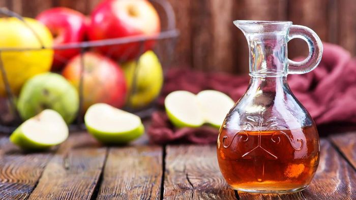 a bottle of apple cider vinegar in the foreground with a heap of apples behind it