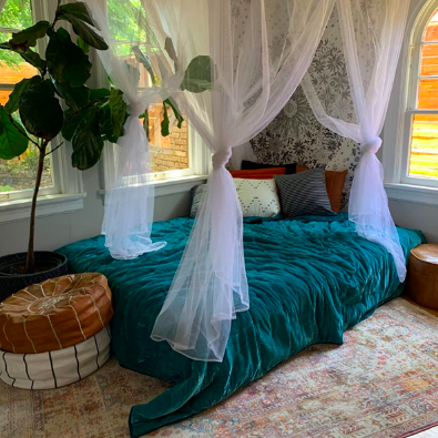 A reviewer captures an image of a mattress tucked in the corner of the room. The bed exudes a well-arranged vibe with floor pillows at both ends, luxurious bedding, and a canopy draped gracefully from the ceiling.