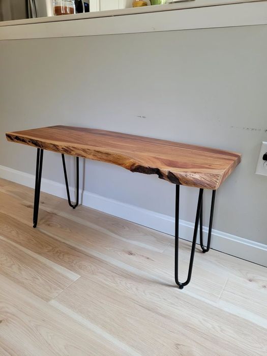 A wooden bench with a live edge top, supported by black metal hairpin legs, set against a light-colored wall.