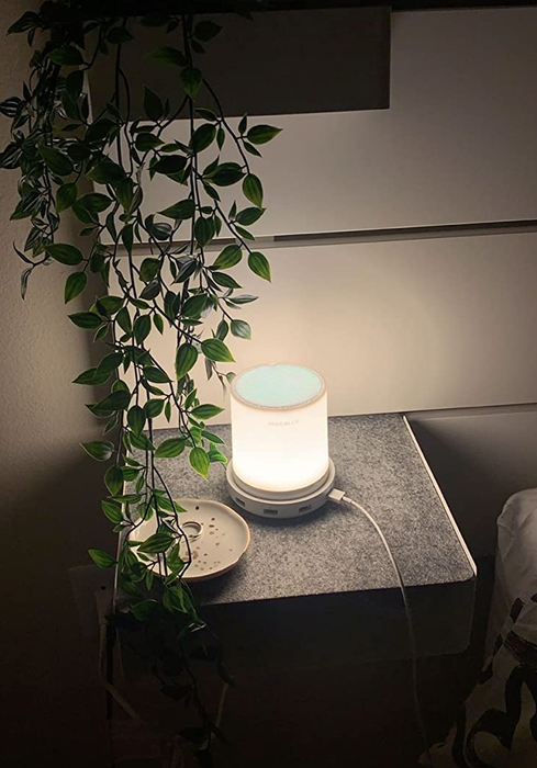 The LED touch lamp shines softly in a dark bedroom, emitting a warm glow, with a cord plugged into the side.