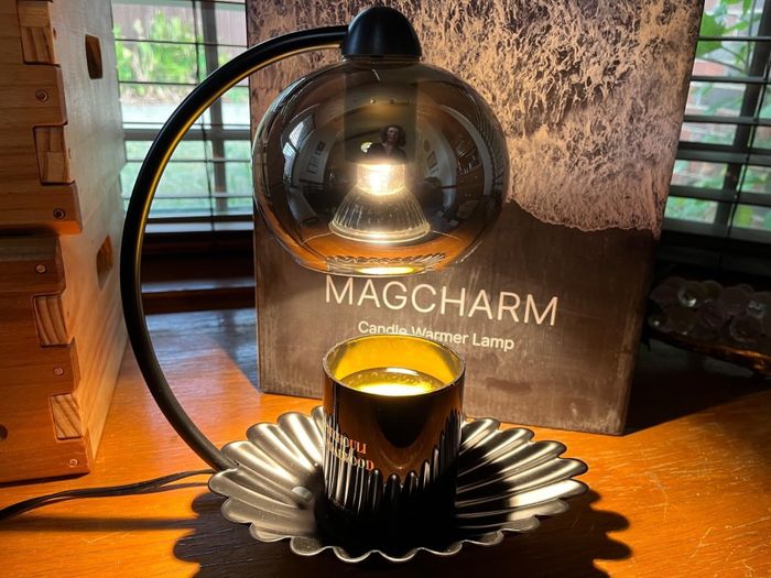 A candle warmer lamp resting on a table beside its box, featuring a modern dome-shaped design with a lit candle underneath