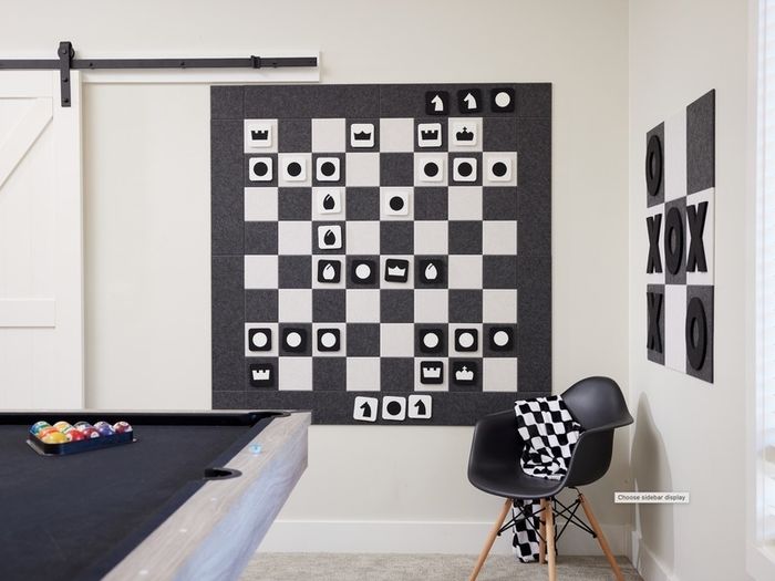 A chic game room features a wall-mounted chess board with large geometric felt pieces. The room also includes a black chair draped with a checkered blanket, a pool table, and a tic-tac-toe wall game.