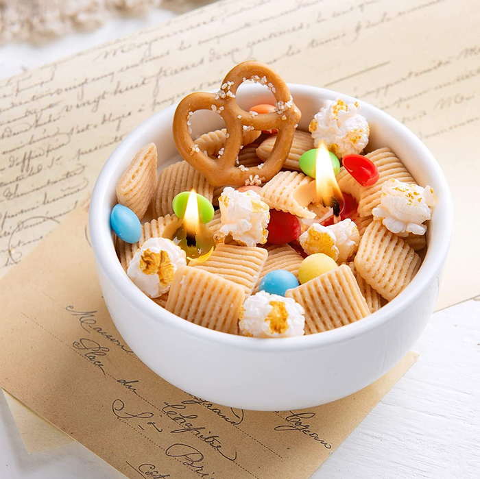 The lit trail mix candle with Chex, popcorn, and pretzels in a small round white bowl.
