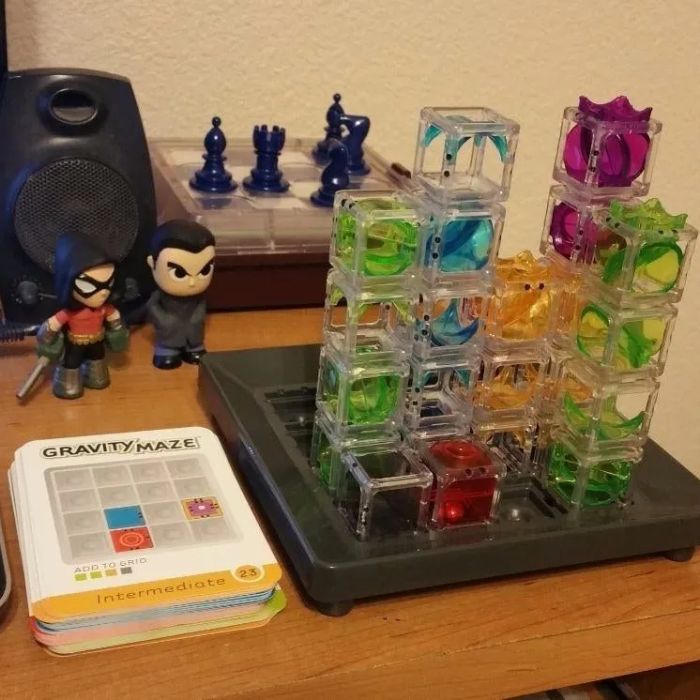 Puzzle game with transparent towers on a table next to action figures and a Gravity Maze card deck. Chess pieces and a speaker are visible in the background