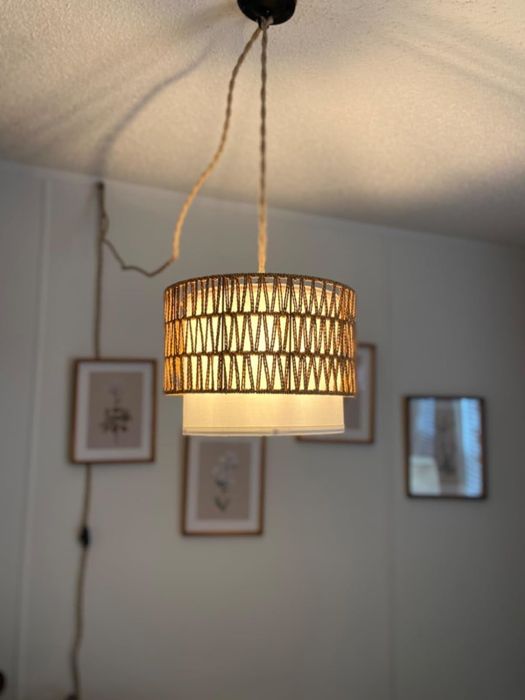A woven pendant light featuring a white inner shade and a natural twine outer shade dangles from the ceiling in a room with framed artwork on the walls, adding a cozy, organic touch to the space.
