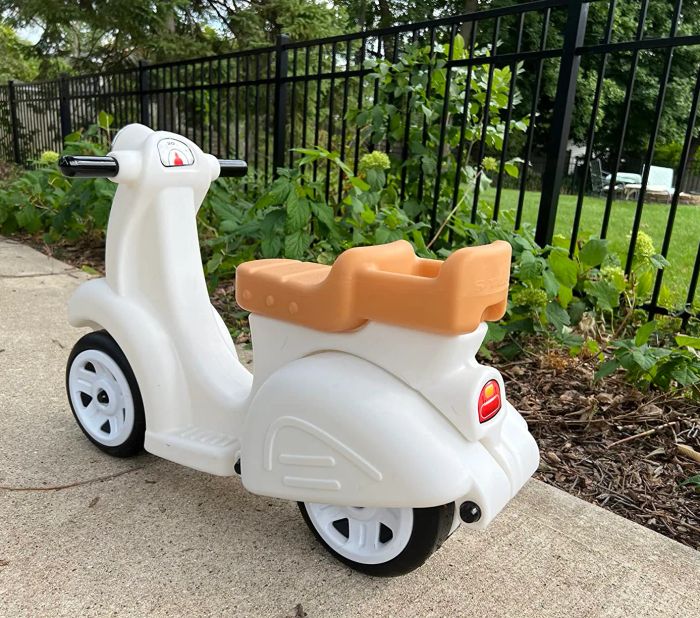 A white and orange toy scooter with three wheels, positioned on a sidewalk next to a garden filled with vibrant greenery.