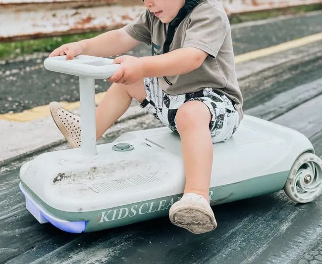 A child wearing patterned shorts riding a kids' cleaning vehicle outdoors